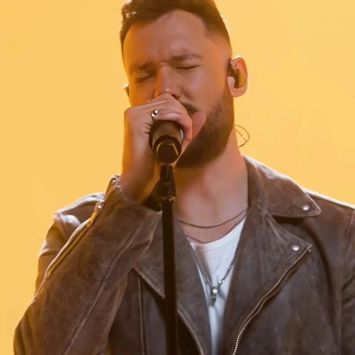 A man in a leather jacket sings into a microphone, wearing a necklace and ring, against a yellow background.
