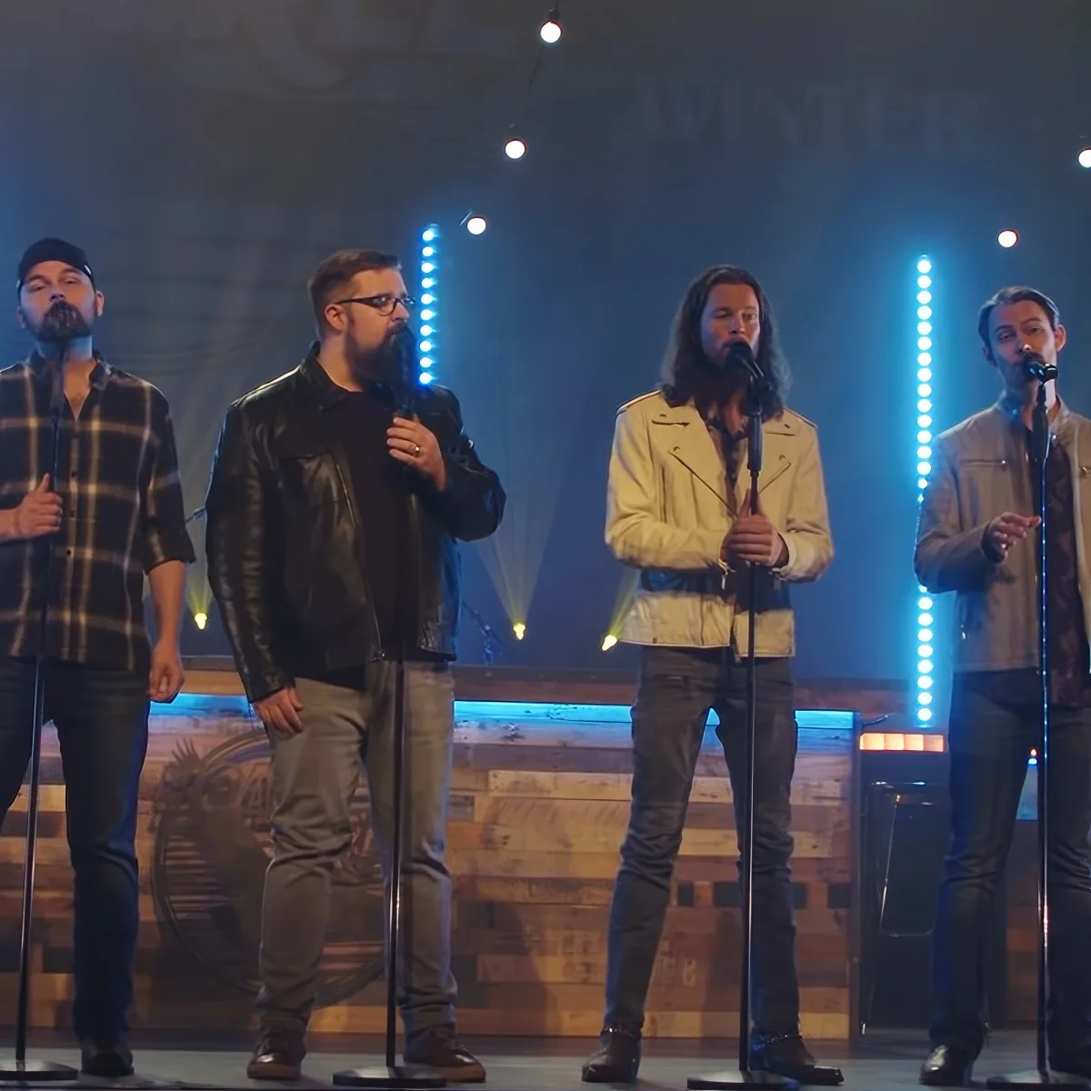 Four men sing on stage with microphones, wearing casual and leather jackets, under blue stage lights.