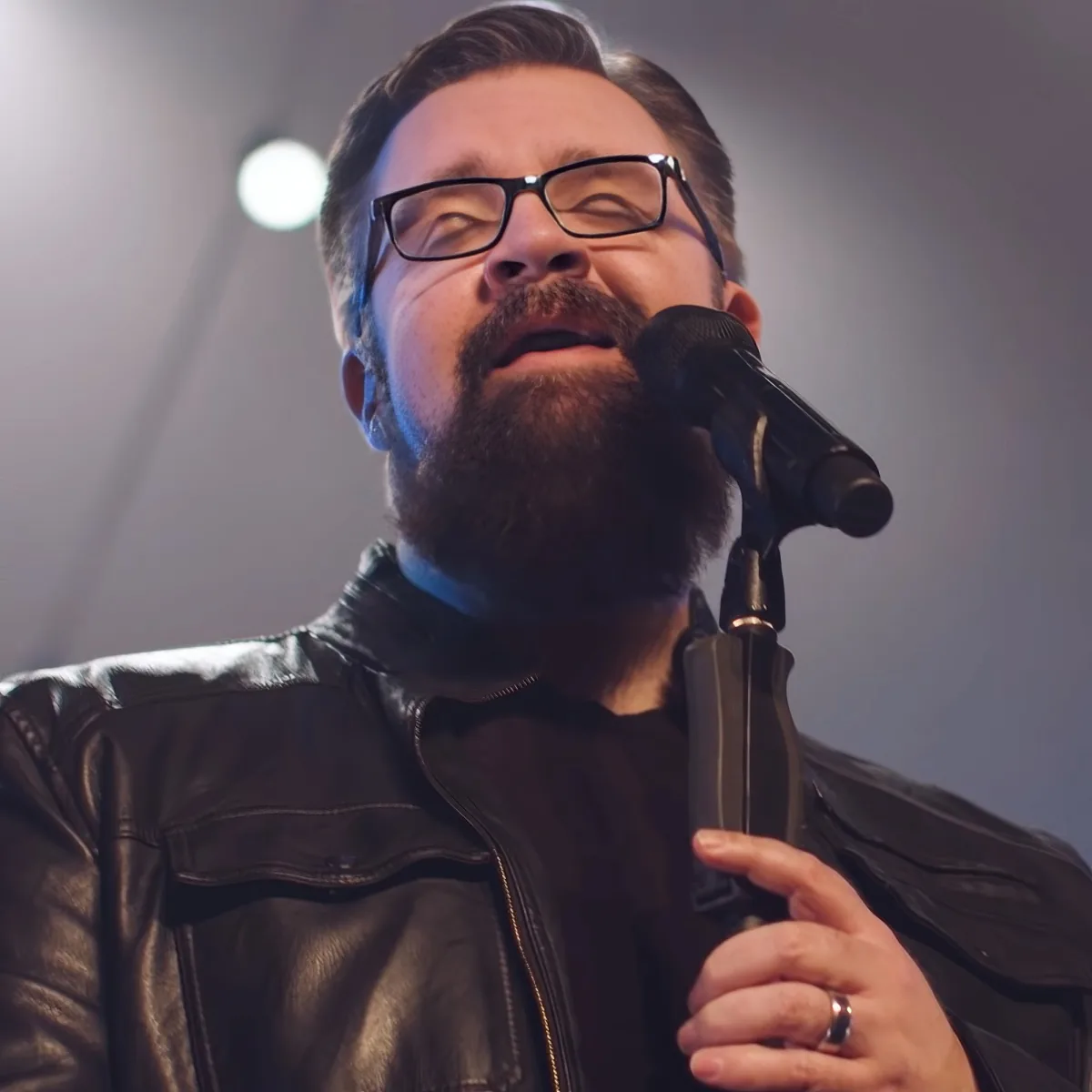 A man with a beard and glasses, wearing a leather jacket, sings into a microphone.