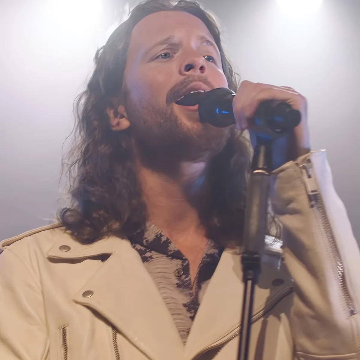 A man with long hair and a beard sings into a microphone, wearing a beige jacket and patterned shirt.
