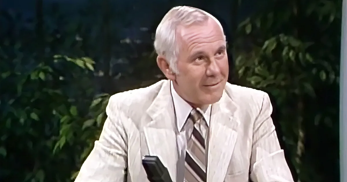 An older man in a white suit and striped tie is seated, smiling, with a microphone in front of him.
