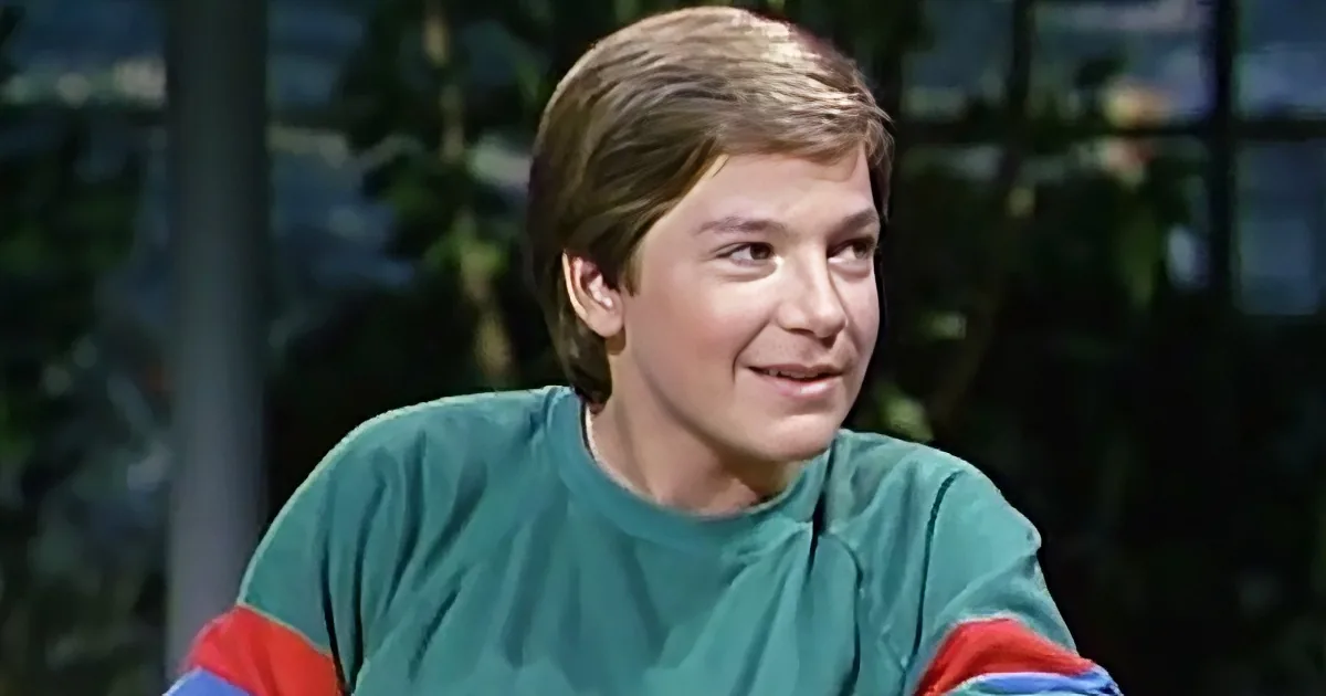 A young person with short brown hair, wearing a green shirt with red and blue stripes, is smiling and looking to the side.