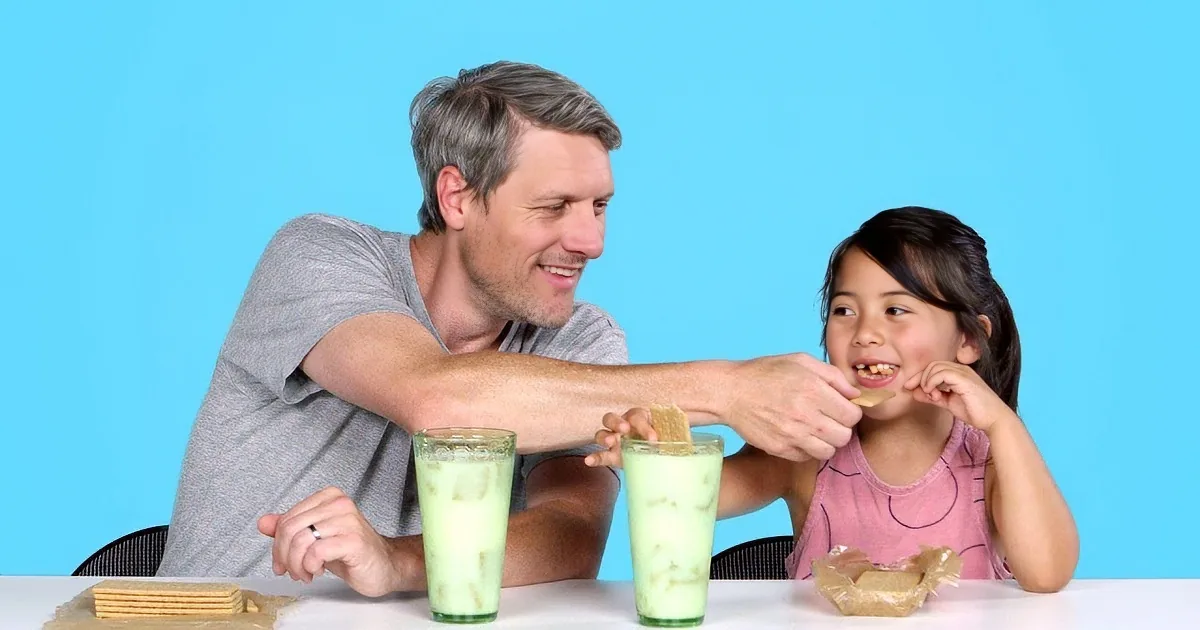 A woman and a boy sit at a table, each holding a sandwich and eating, with bowls of cereal in front of them.
