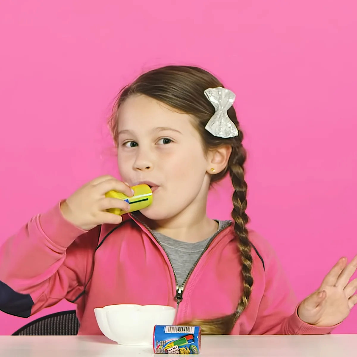 A girl with braids and a bow in her hair drinks from a yellow cup while sitting at a table with a bowl and a box.