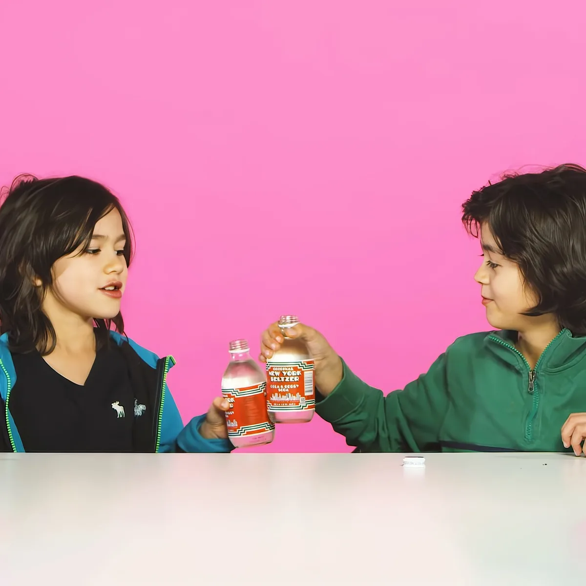 Two children hold up bottles of New York Seltzer in front of a pink background.