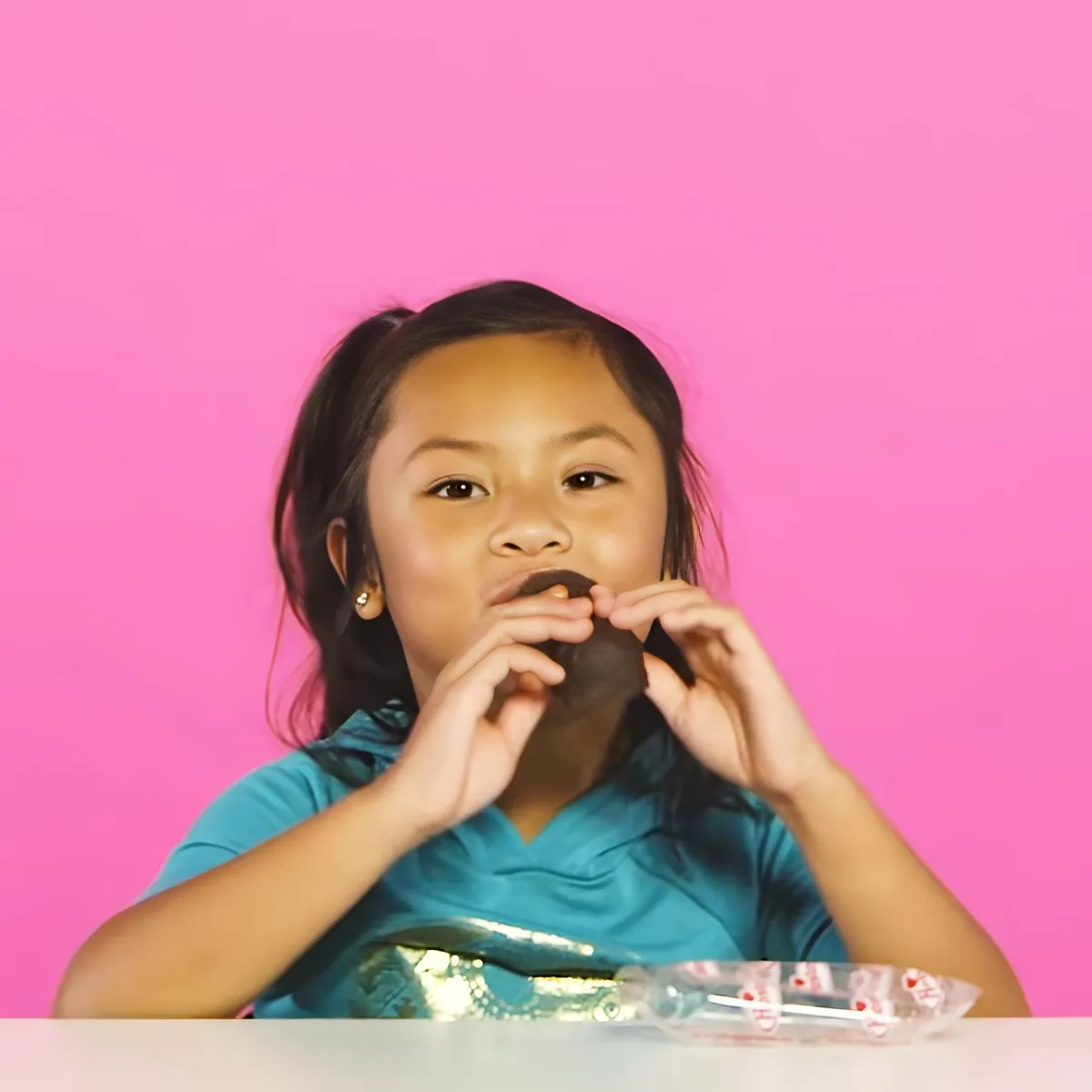 A young girl in a teal shirt holds a chocolate bar, with a pink background.