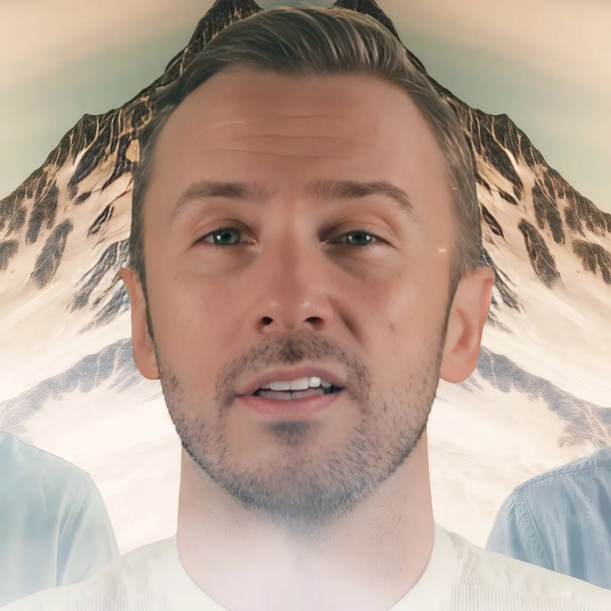 A man with short hair and a beard, wearing a light blue shirt, is in front of a snowy mountain backdrop.