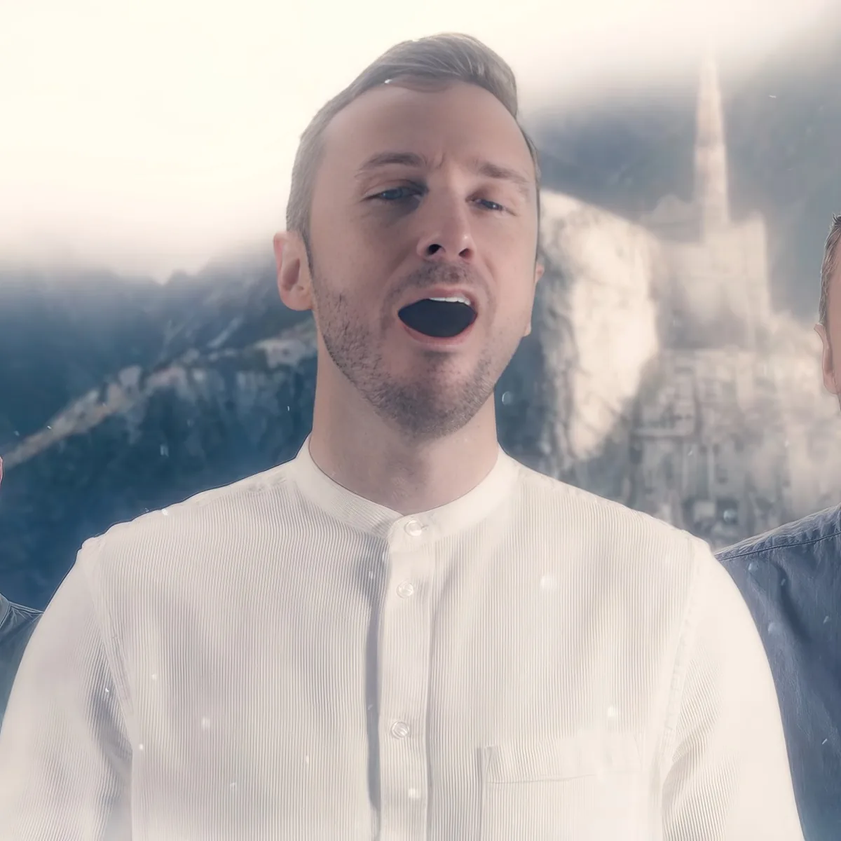 A man in a white shirt sings with a snowy mountain and church steeple in the background.