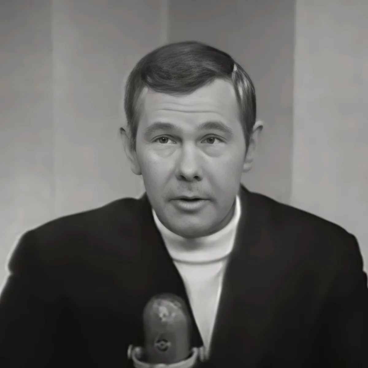 A man in a suit and turtleneck speaks into a microphone, likely in a studio setting.