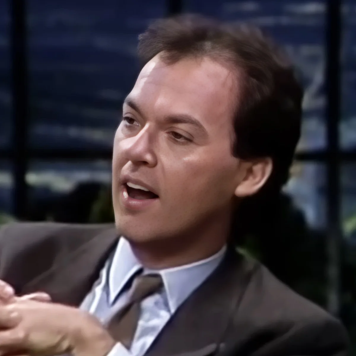 A man in a suit and tie is gesturing with his hands, appearing to speak or explain something.