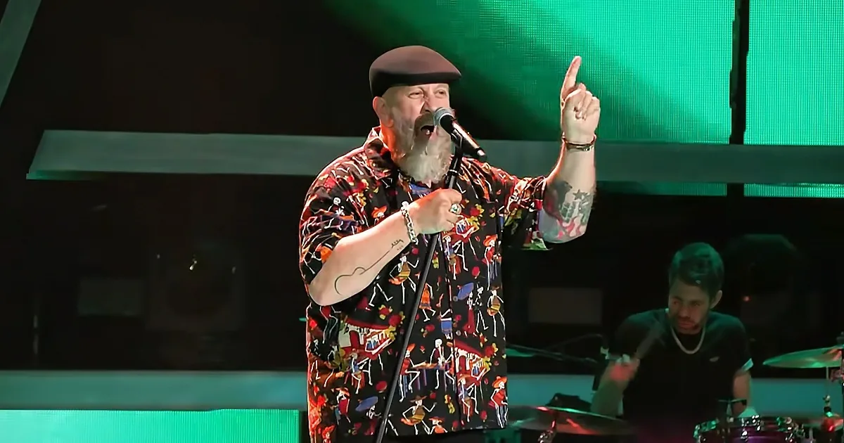 A man in a colorful shirt and beret sings into a microphone, pointing upwards, with a drummer in the background.