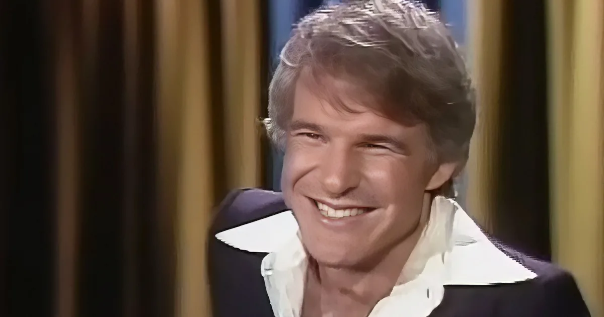 A smiling man with short, light brown hair, wearing a white shirt with a dark jacket, against a blurred background.