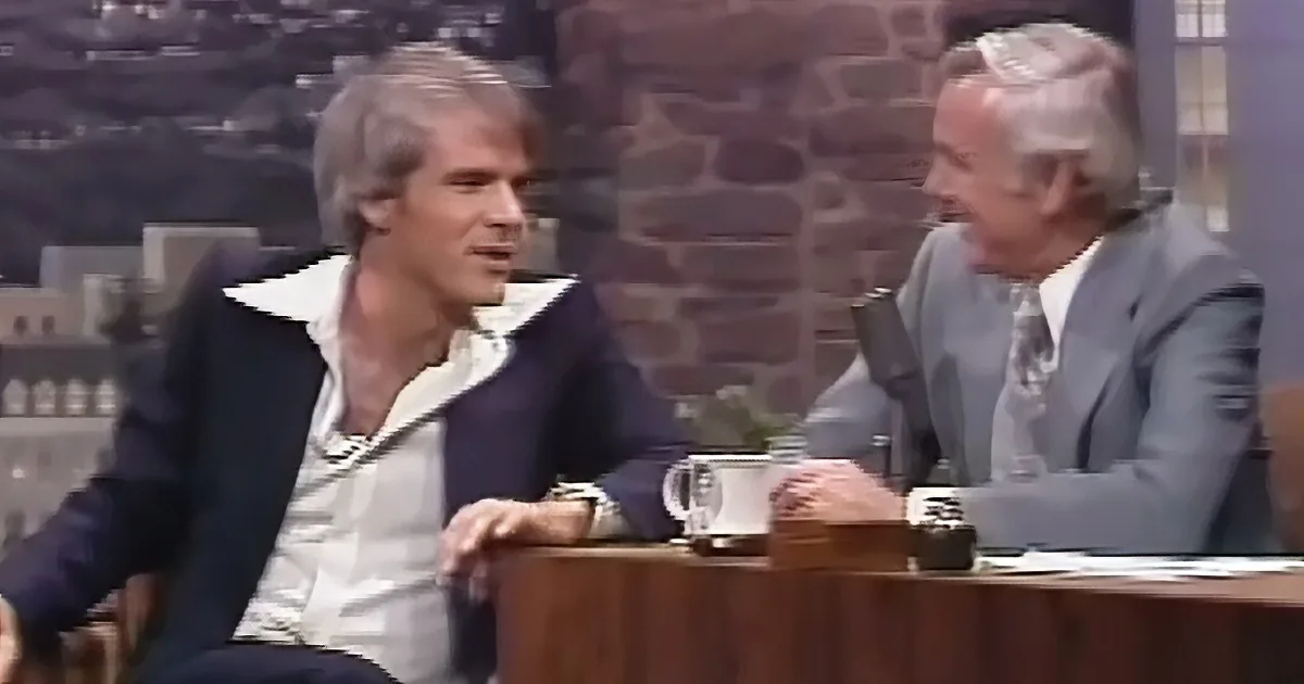 Two men are seated at a table, one in a suit, the other in a casual outfit, engaged in conversation.
