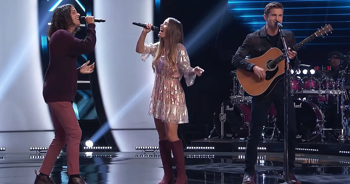 Three performers sing on stage, one with a guitar, another in a sparkly dress, and the third in a maroon outfit.