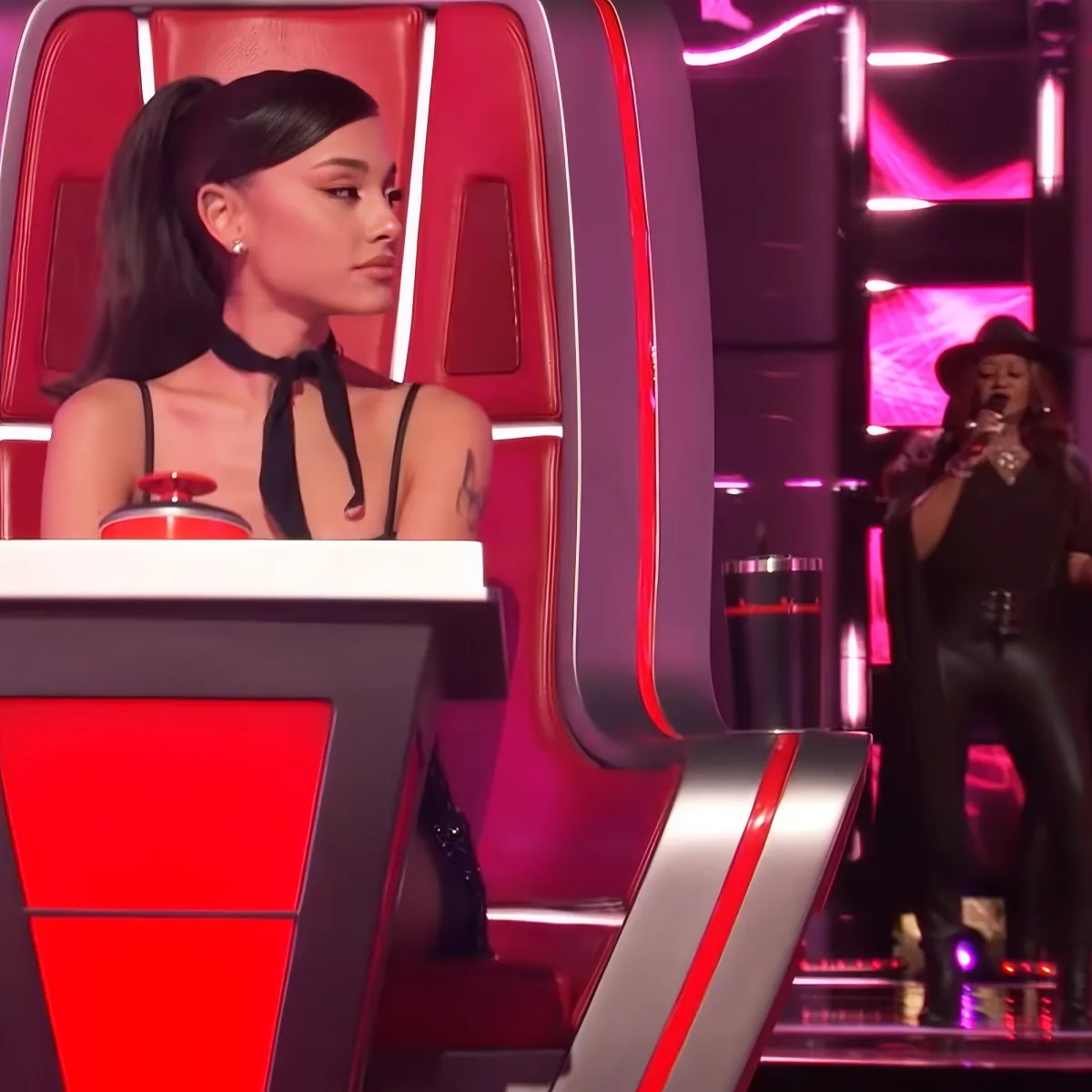 A woman in a black outfit sits on a red chair, watching another woman perform on stage.