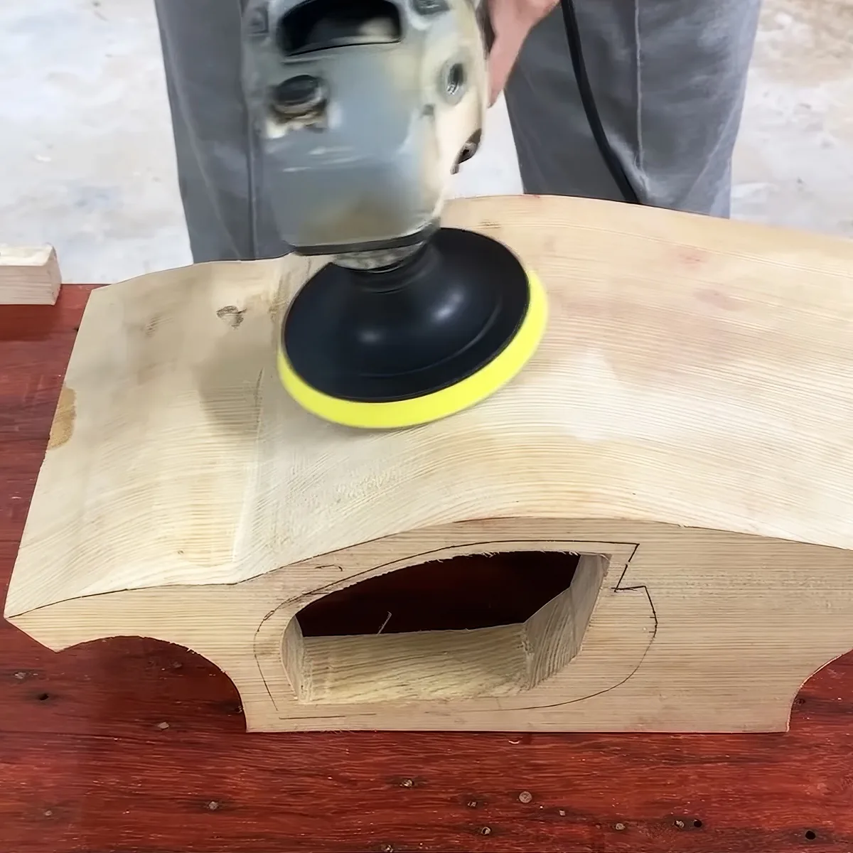 A person uses a sander to smooth a curved wooden piece on a red workbench.
