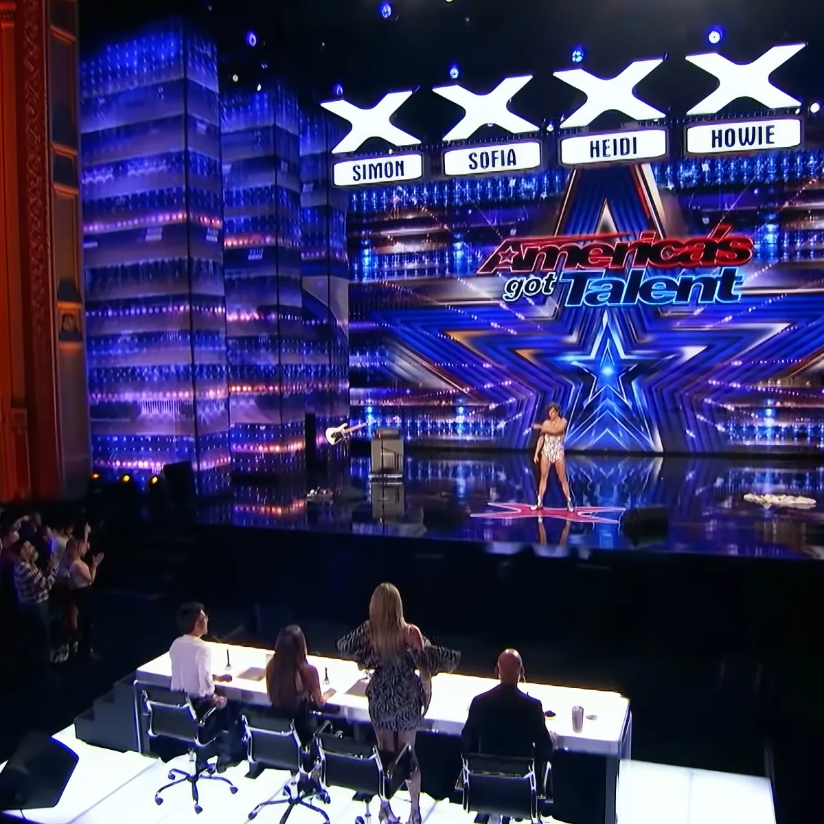 A performer stands on a stage with judges' names displayed above, in front of a large audience.