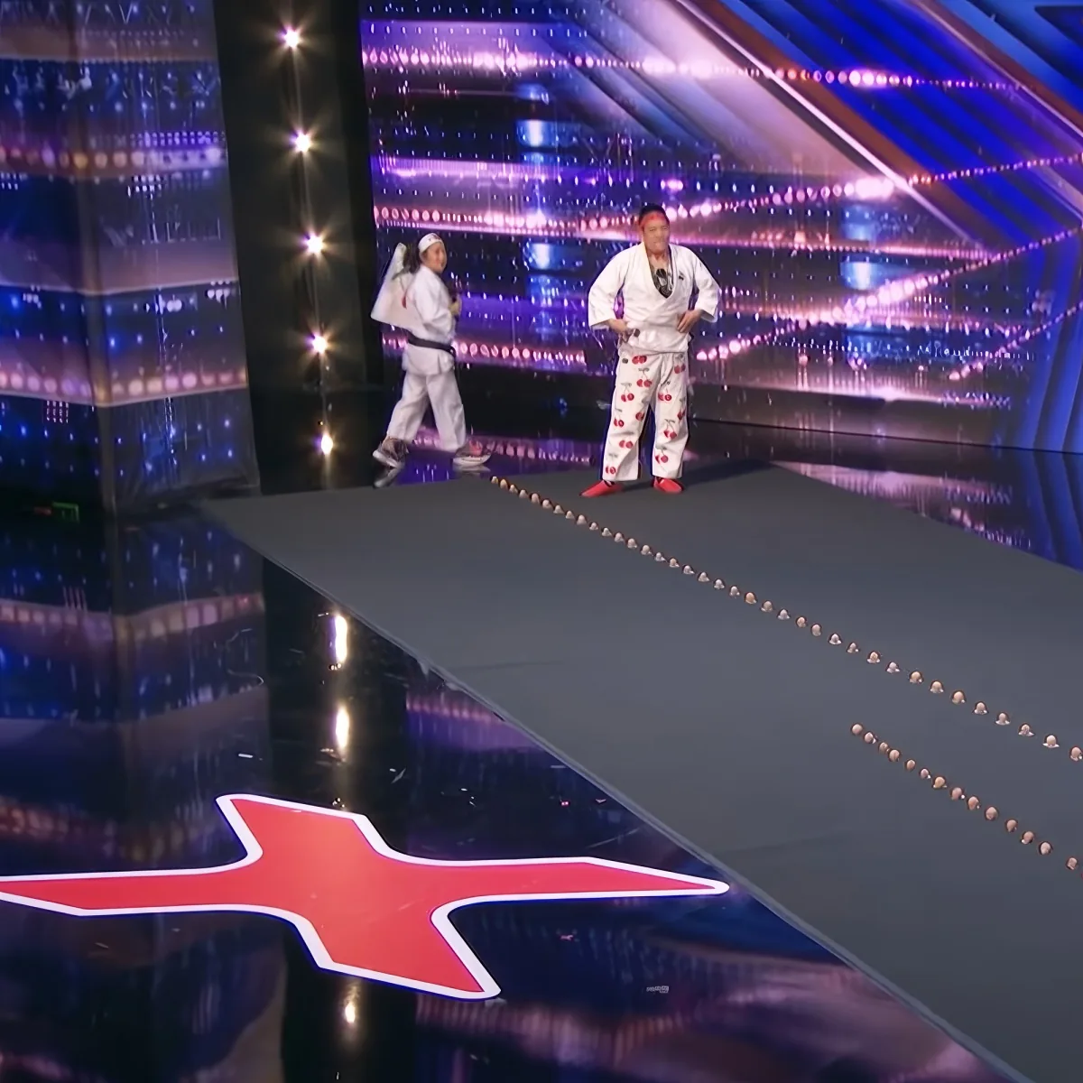 Two people in karate uniforms stand on a stage with a reflective floor and colorful lights.