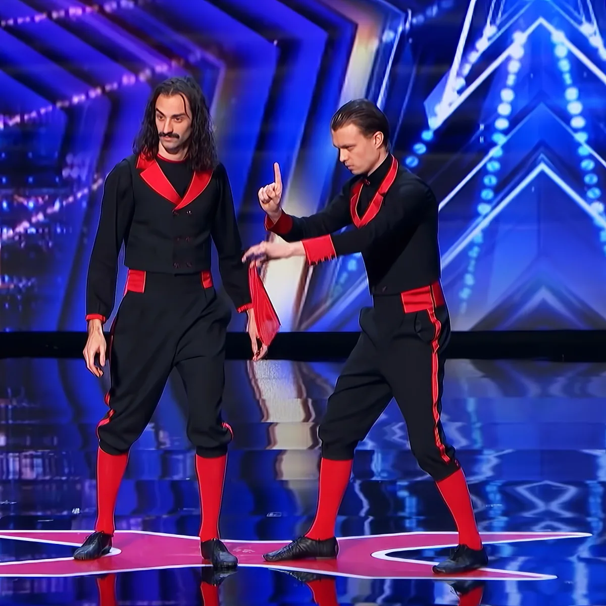 Two performers in black and red outfits dance on a stage with a blue-lit background.