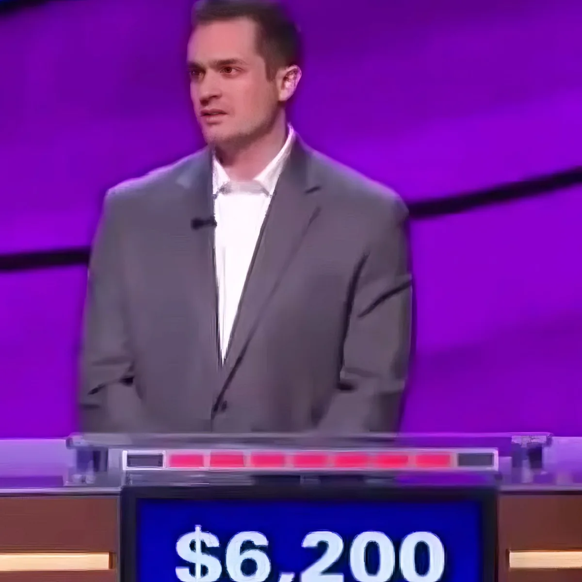 A man in a gray suit stands behind a podium with a score of $6,200 on a game show set.