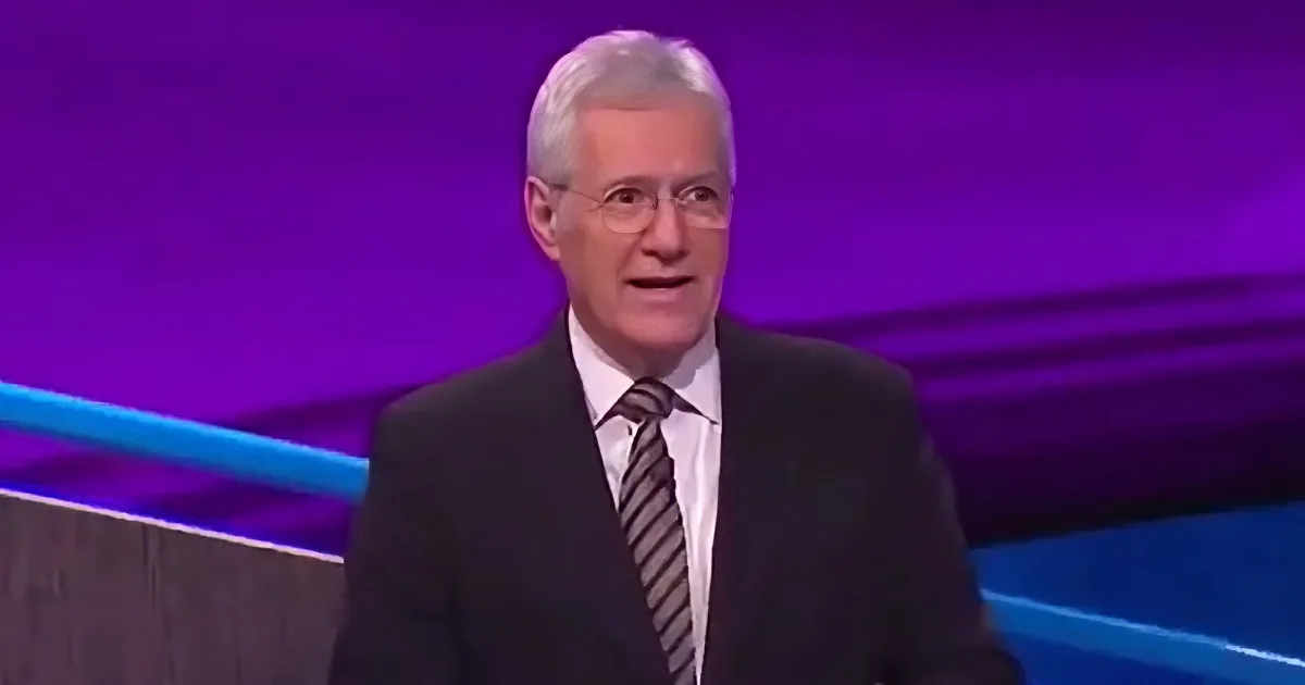 A man in a suit and tie stands on a stage with a purple background.