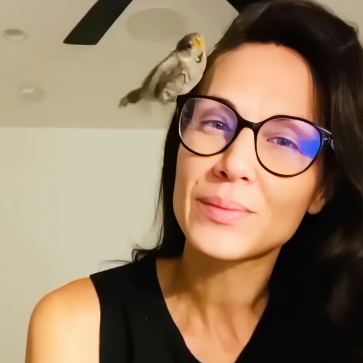 A woman with glasses and a bird perched on her shoulder, both against a white wall.
