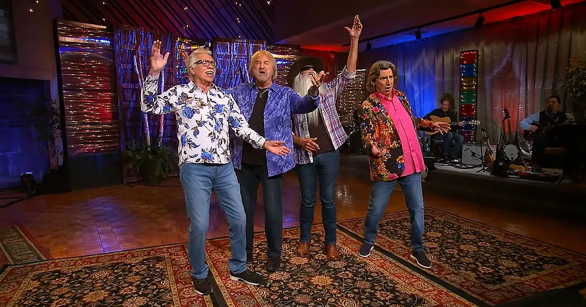 Four people in vibrant clothing are singing and dancing on a stage with musicians in the background.