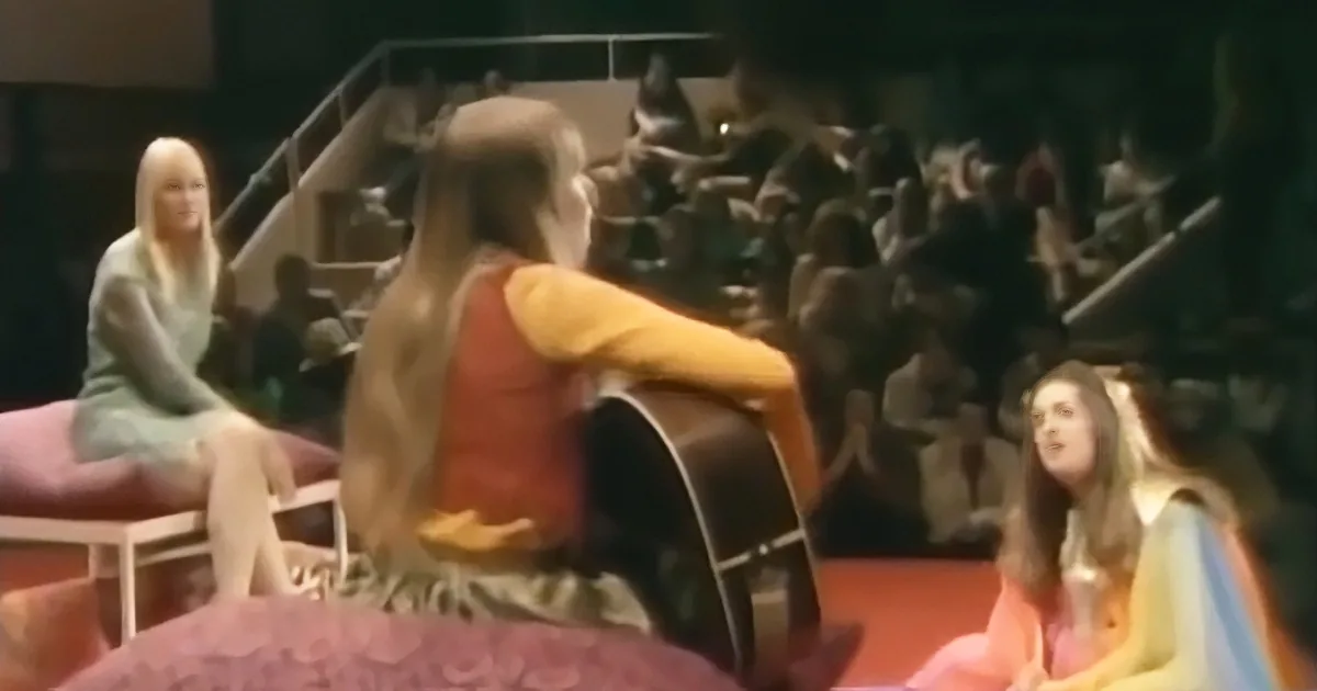 A person with long hair plays guitar on stage, surrounded by an audience in a dimly lit venue.
