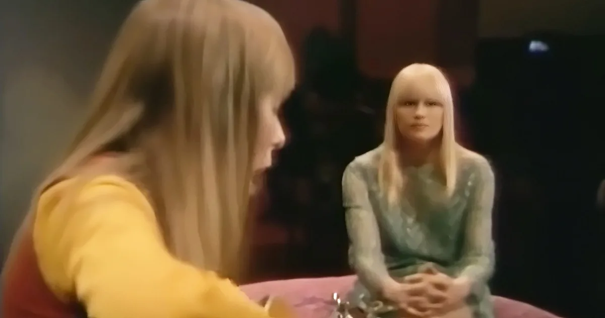 A woman with long blonde hair in a yellow top is facing another woman in a green dress with long blonde hair.