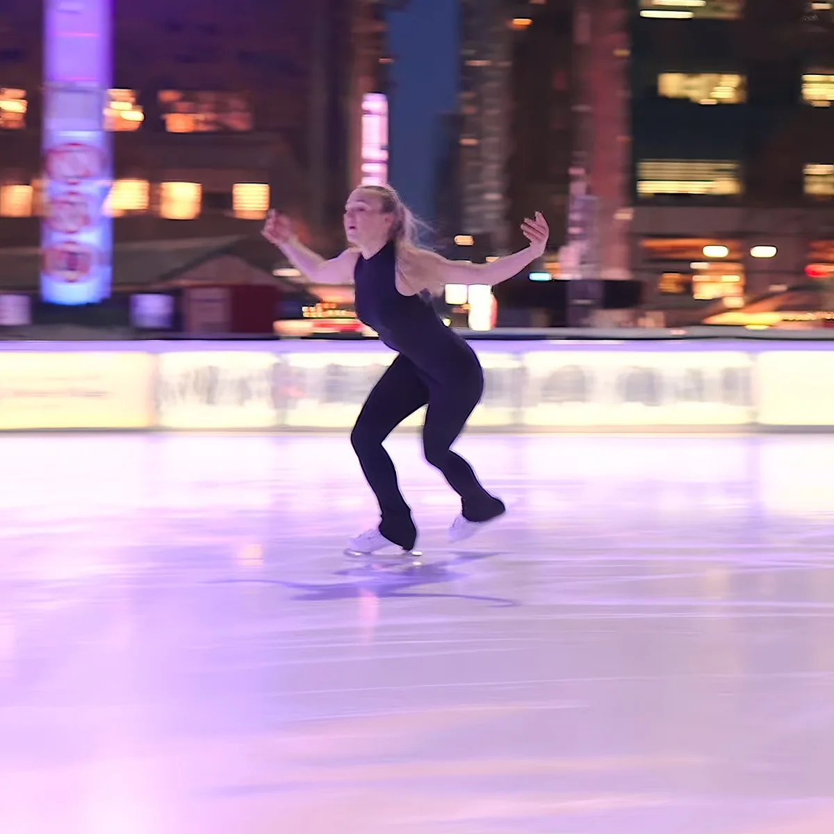 A figure skater in a black outfit glides gracefully on an ice rink with city lights in the background.