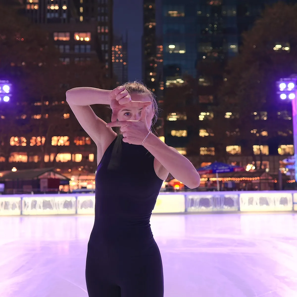 A person in a black outfit makes a frame with their hands on an ice rink with city lights in the background.