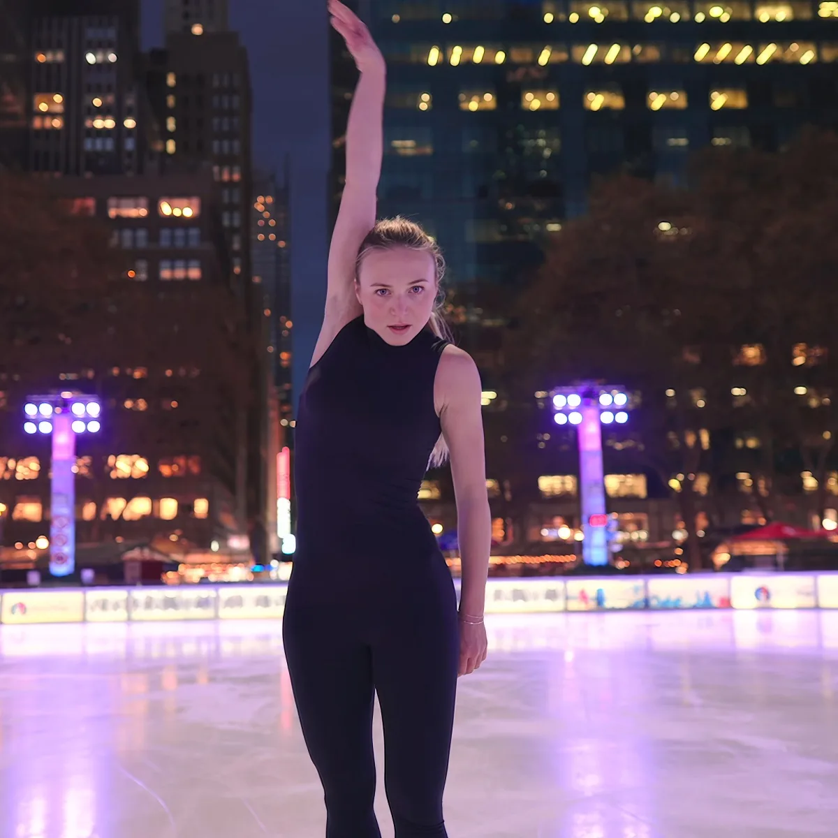 A person in a black outfit stands on an ice rink with arms raised, city lights glowing in the background.