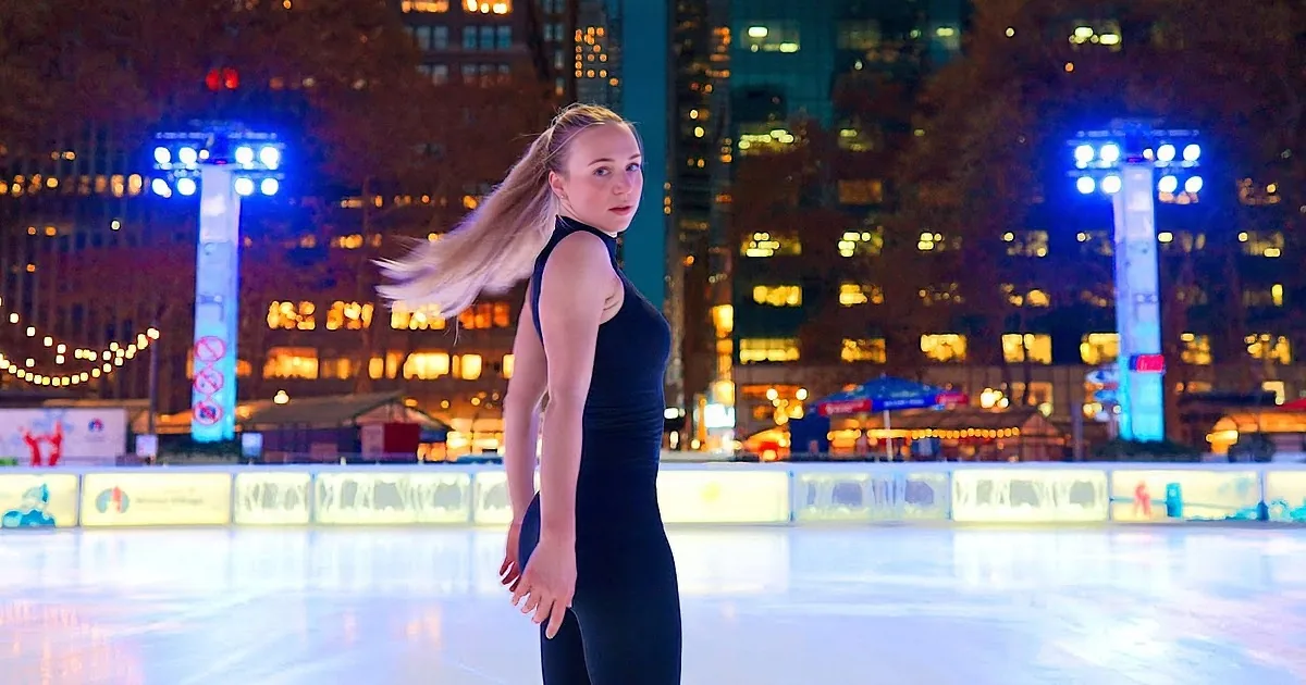 A woman in a black outfit skates gracefully on an ice rink at night, with city lights and umbrellas in the background.
