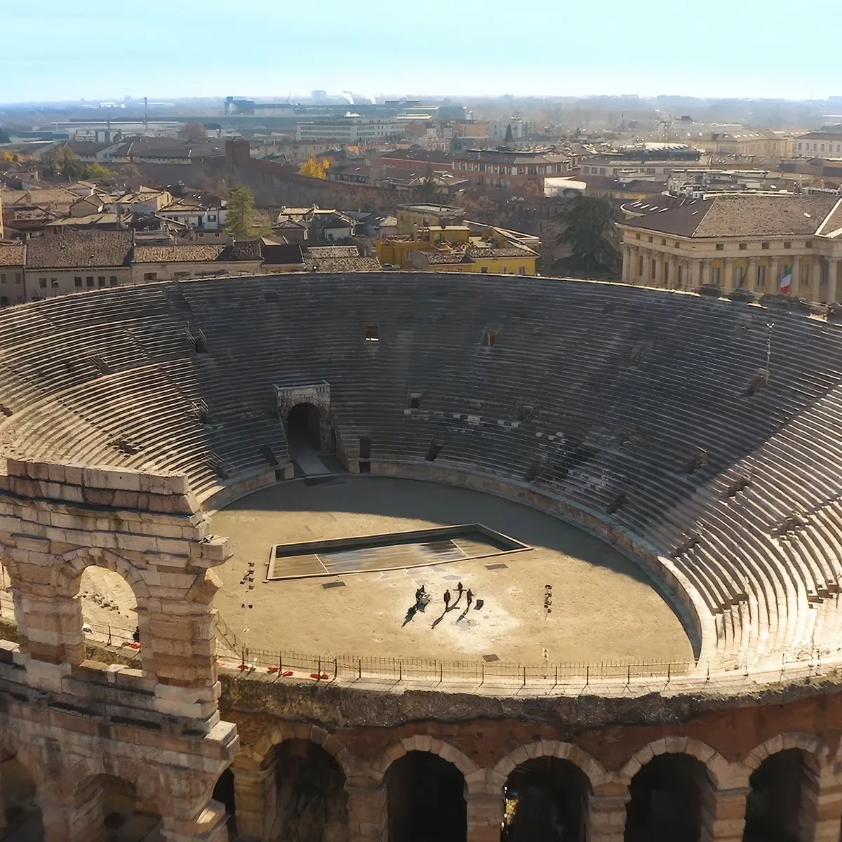 An ancient Roman amphitheater with tiered seating and a central arena, surrounded by modern buildings in the background.