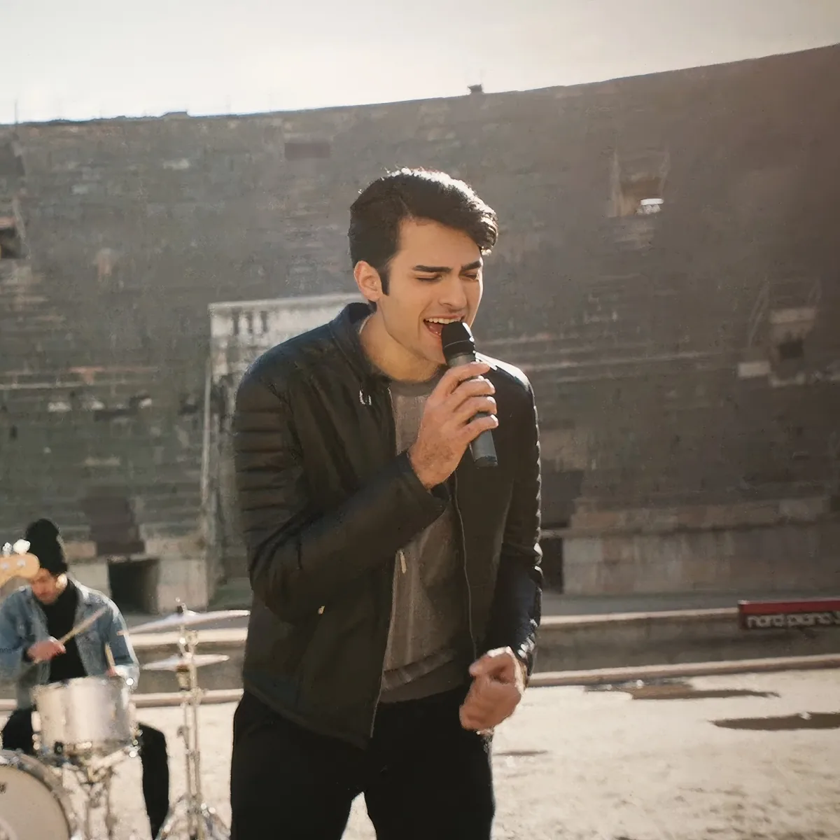 A man in a black jacket sings into a microphone in front of an ancient amphitheater.