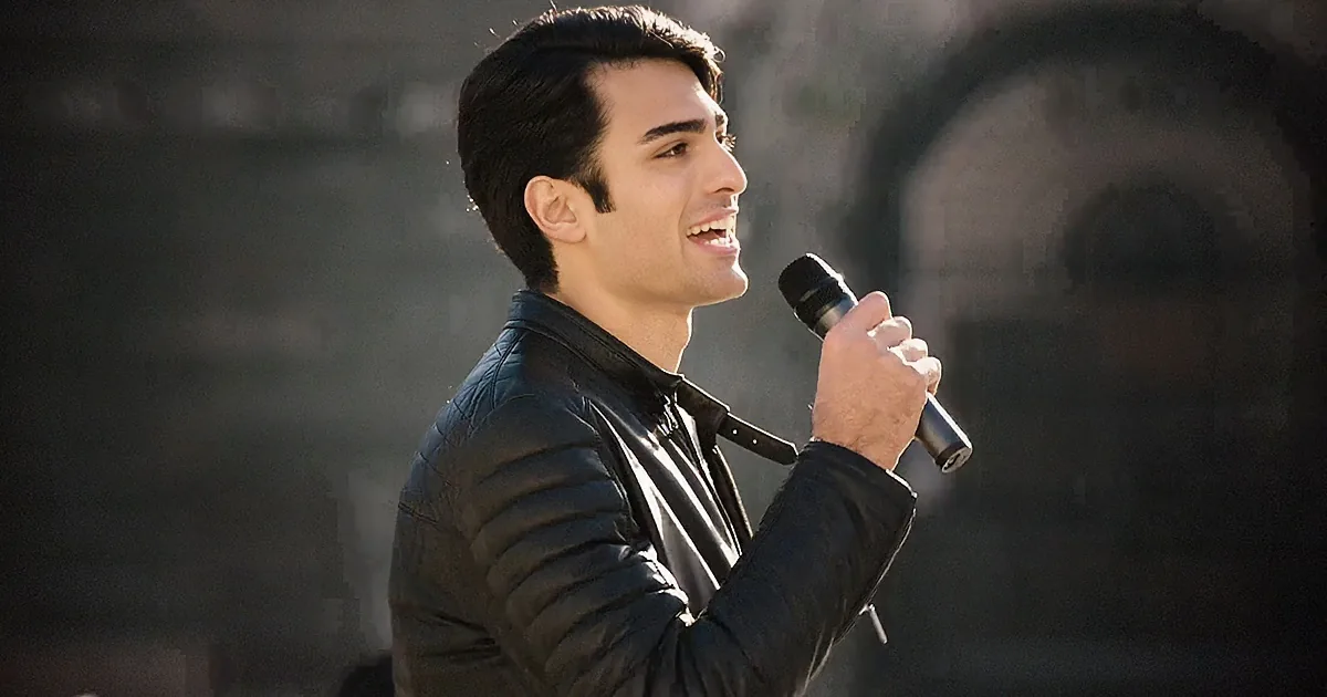 A man in a black leather jacket holds a microphone, smiling, with a blurred background.