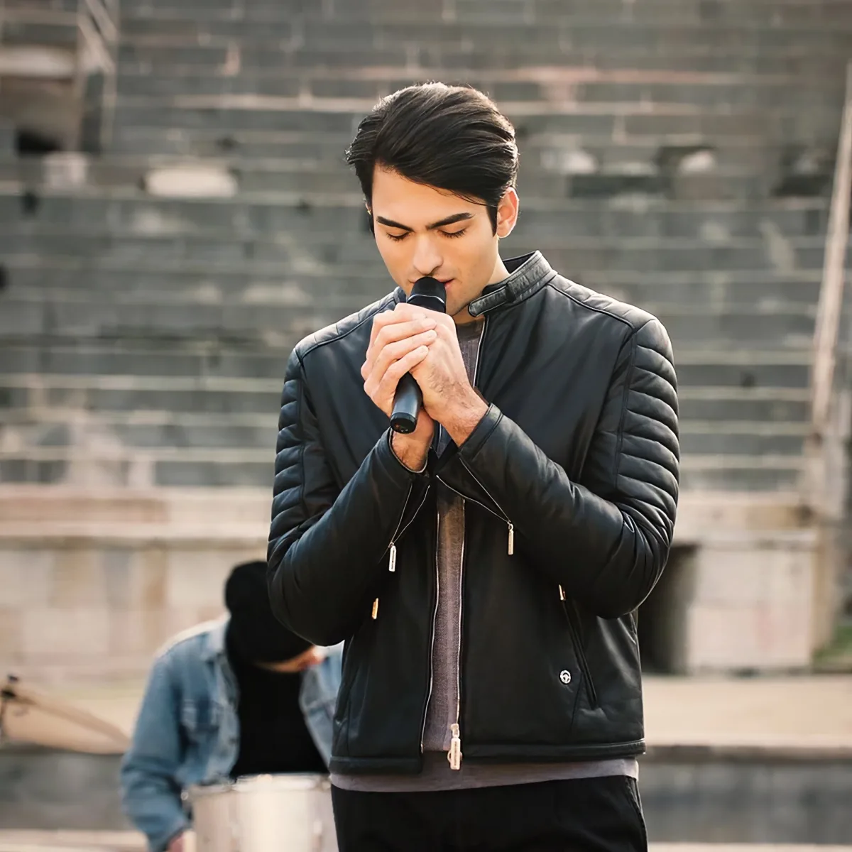 A man in a black leather jacket holds a microphone, singing passionately in front of a brick wall.