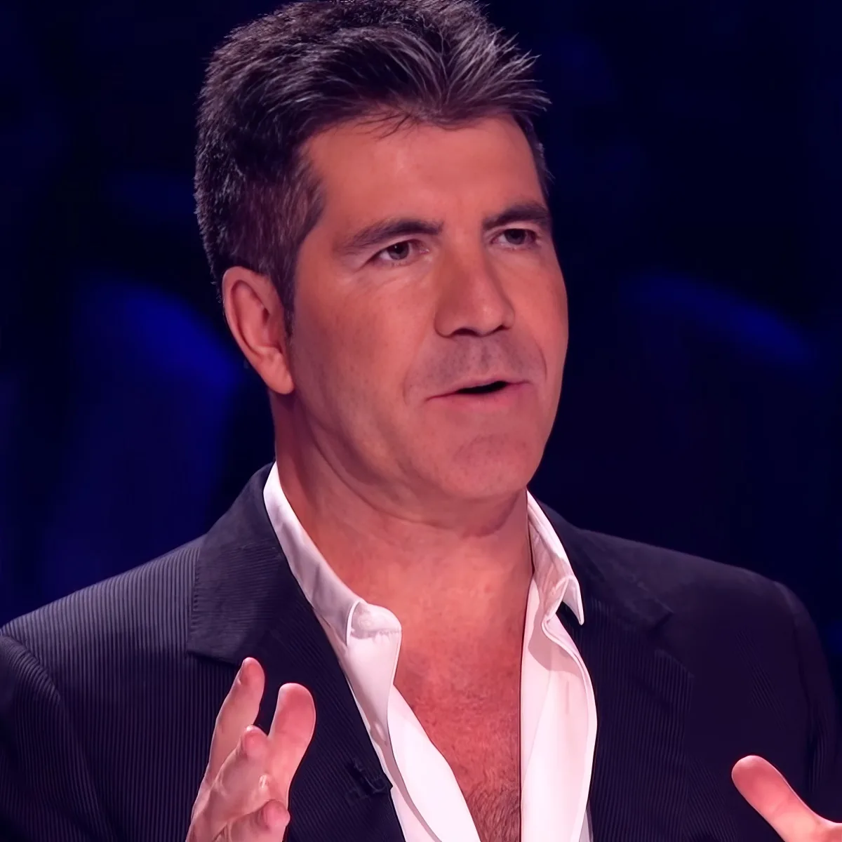 A man in a suit and open shirt gestures with his hands while speaking.