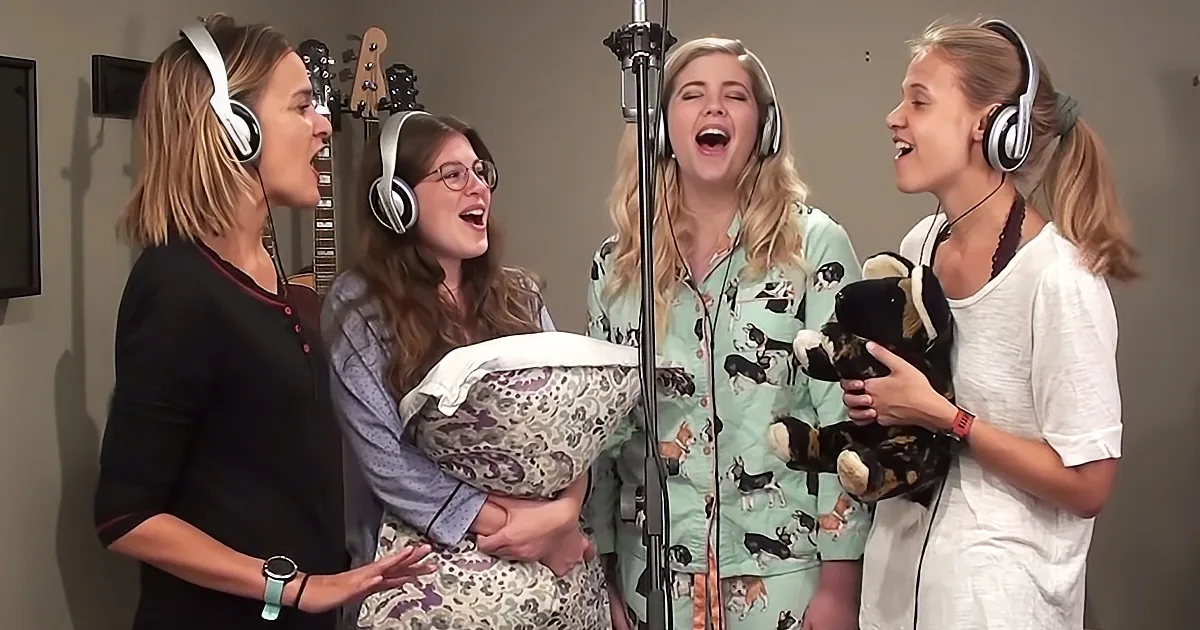 Four women in a recording studio, wearing headphones, singing and holding stuffed animals.