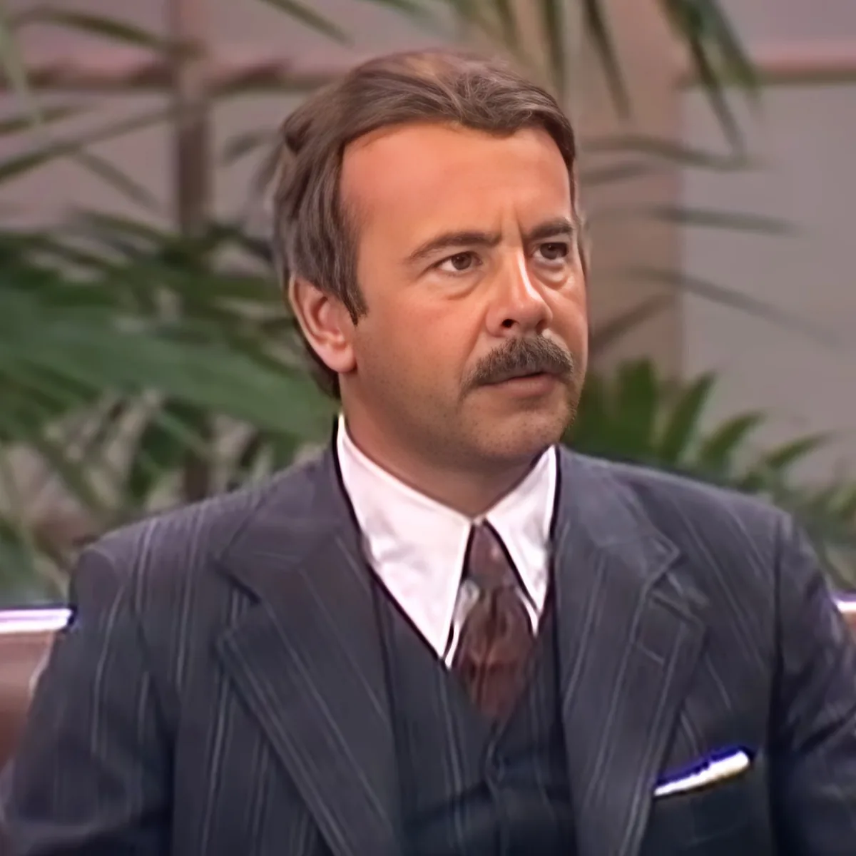 A man in a pinstriped suit and tie, holding a pen, appears to be in a formal setting with plants in the background.