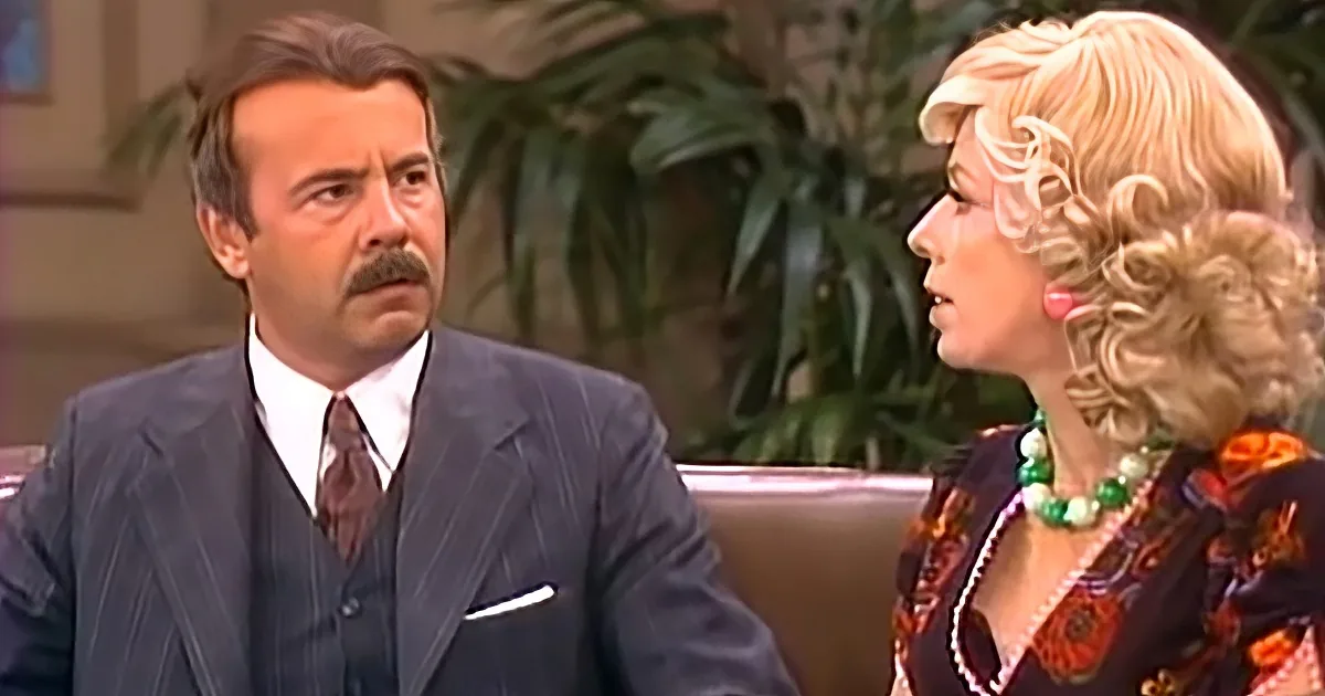 A man in a suit and tie sits next to a woman with blonde hair and a colorful outfit, both appearing to be in a conversation.