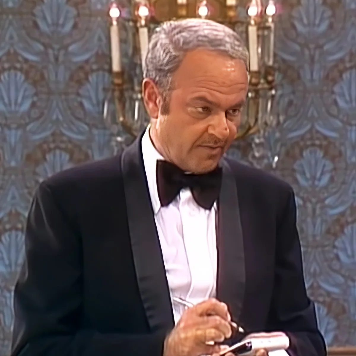 A man in a tuxedo holding a pen, wearing a bow tie, with a patterned wall and chandelier in the background.