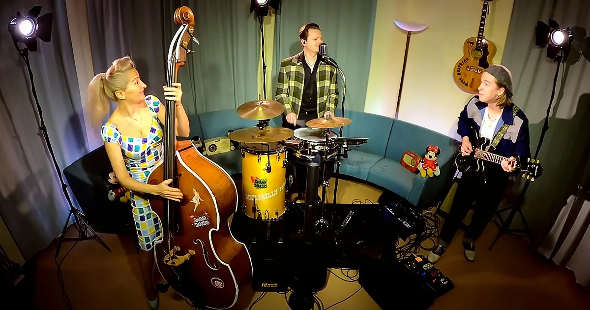 A woman plays a double bass in a small studio with a drummer and guitarist.