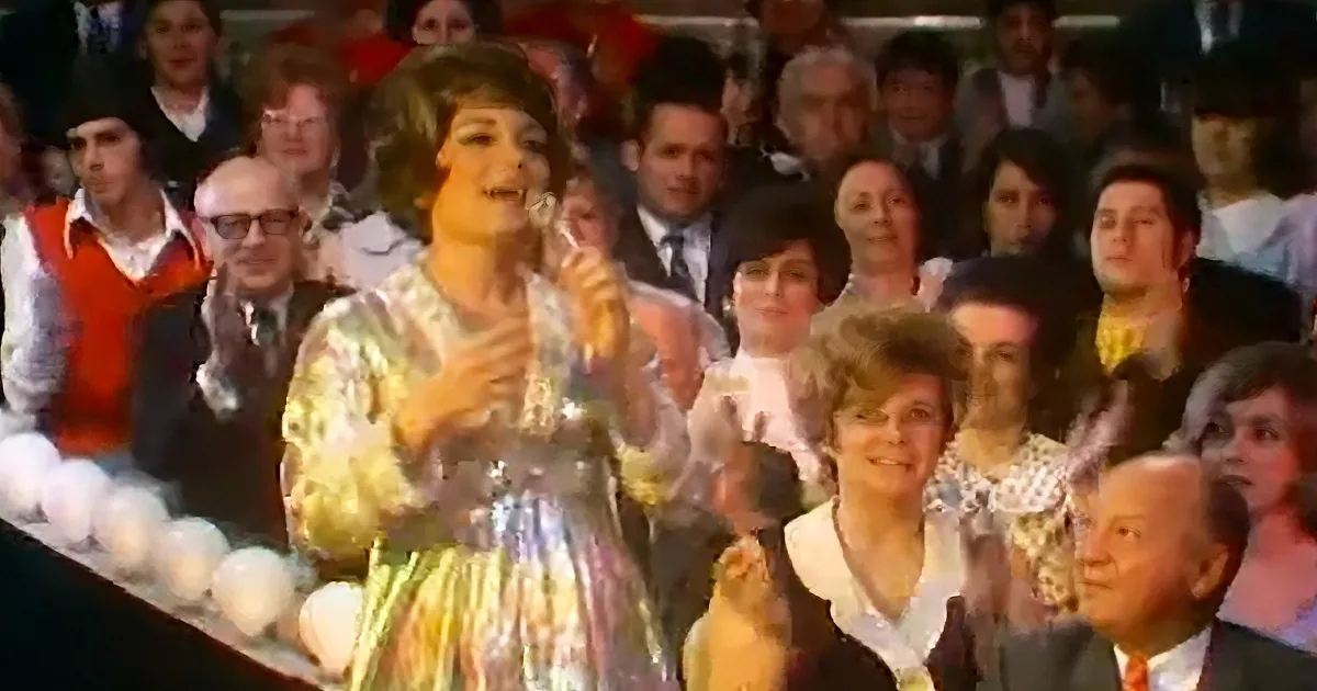 A woman in a colorful dress sings into a microphone, surrounded by an audience clapping.