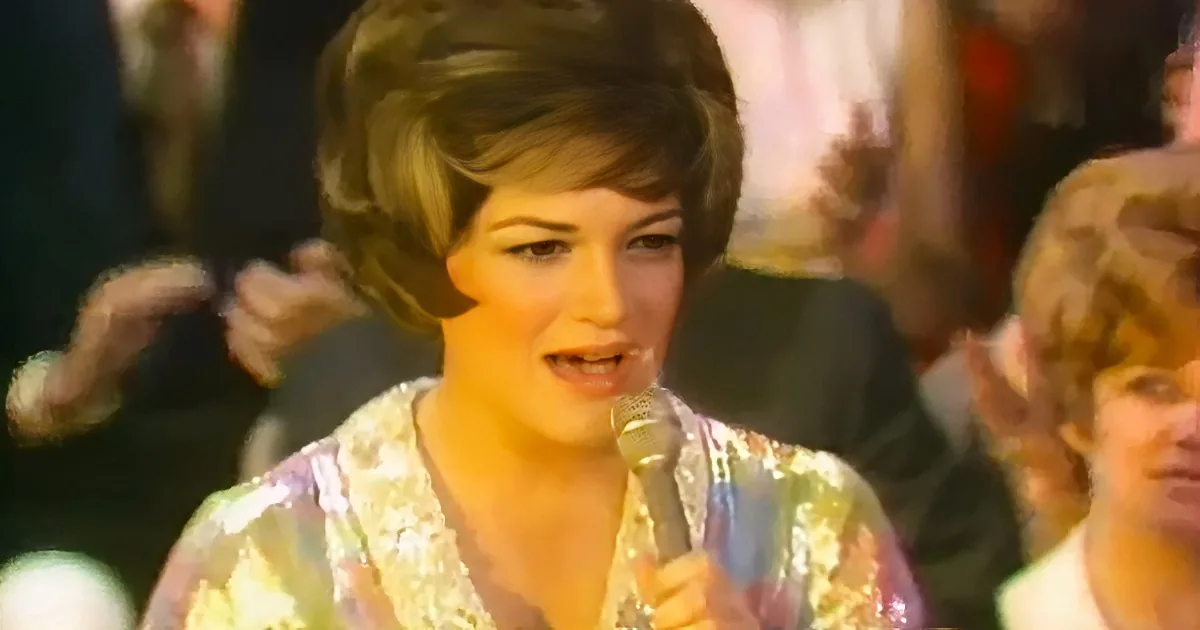 A woman with short, curly hair sings into a microphone, wearing a sparkly, patterned top, with a crowd in the background.