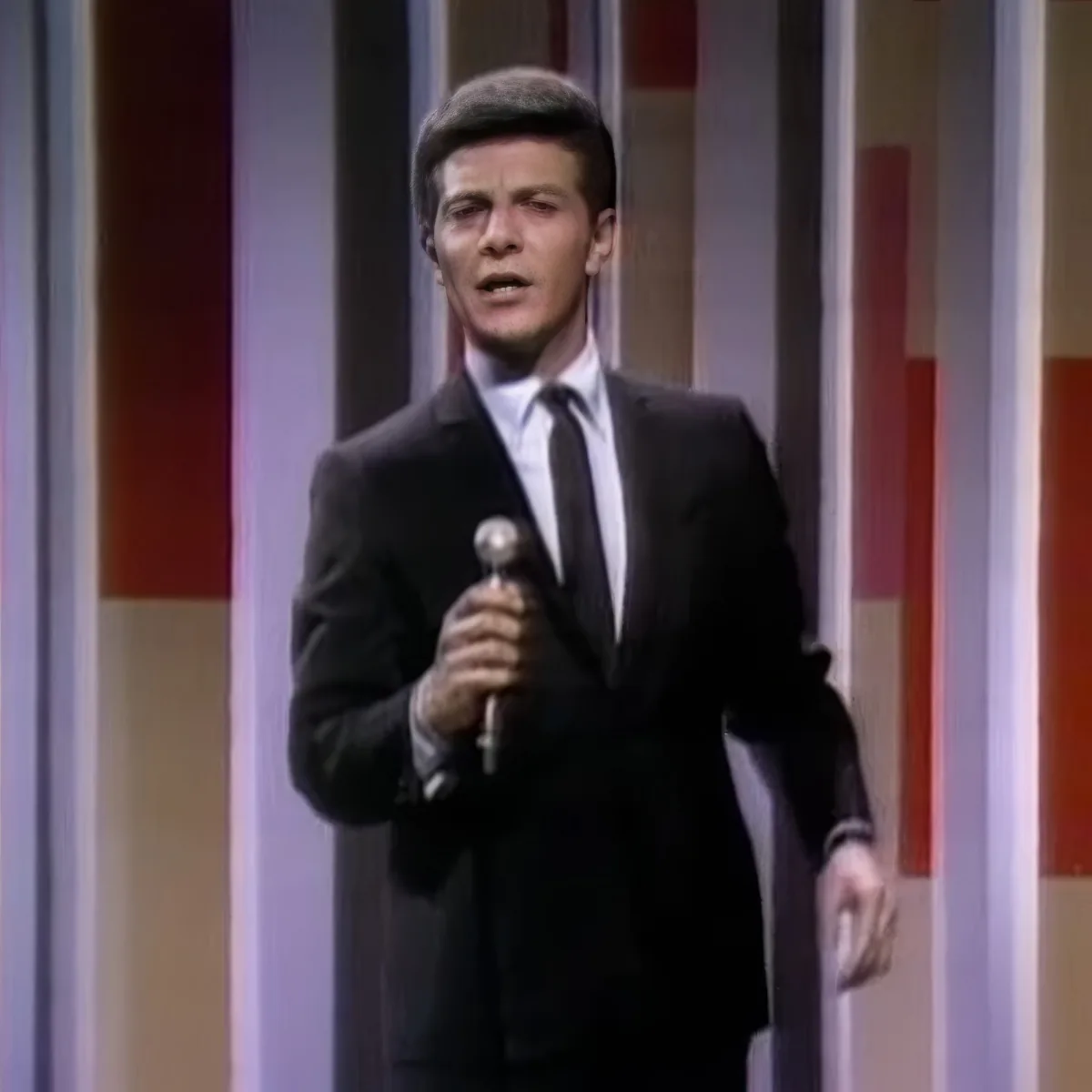 A man in a black suit and tie holds a microphone, standing against a colorful striped background.