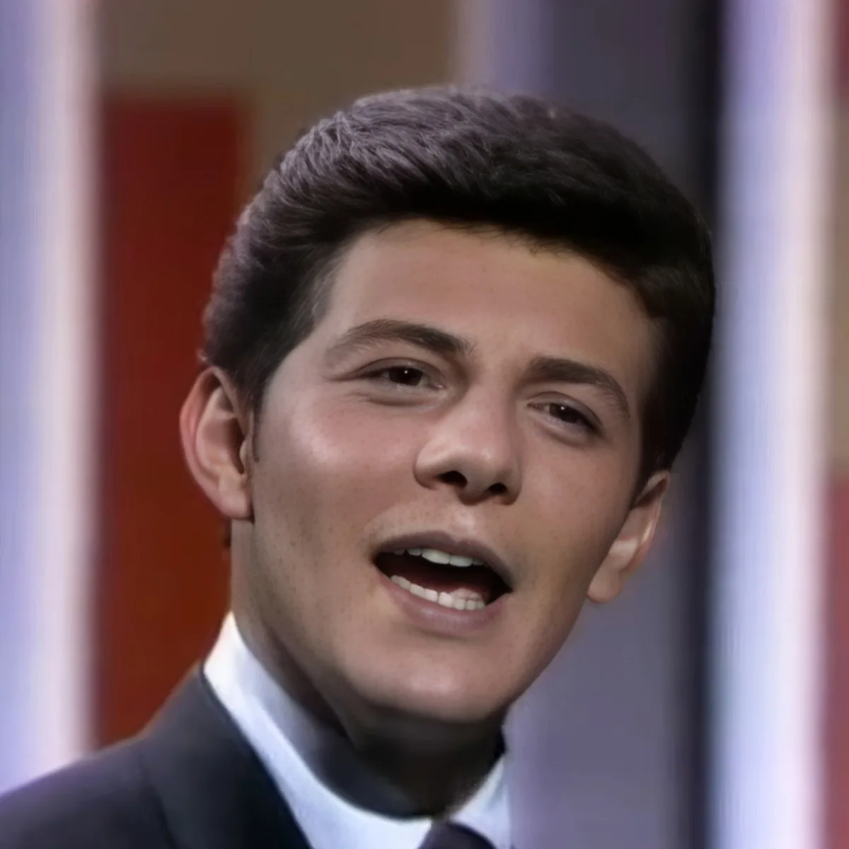 A man with dark hair, wearing a suit, is singing with his mouth open.
