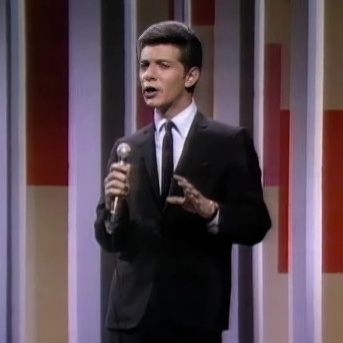 A man in a suit and tie holds a microphone, singing on stage with a colorful striped background.