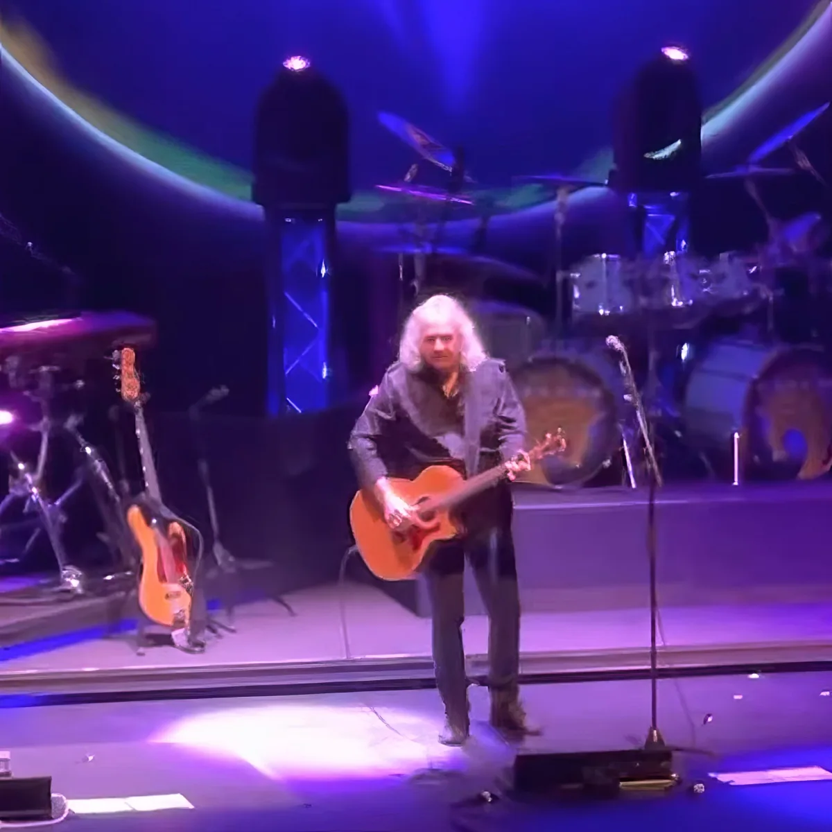 A musician with long hair plays an acoustic guitar on stage under purple lighting.