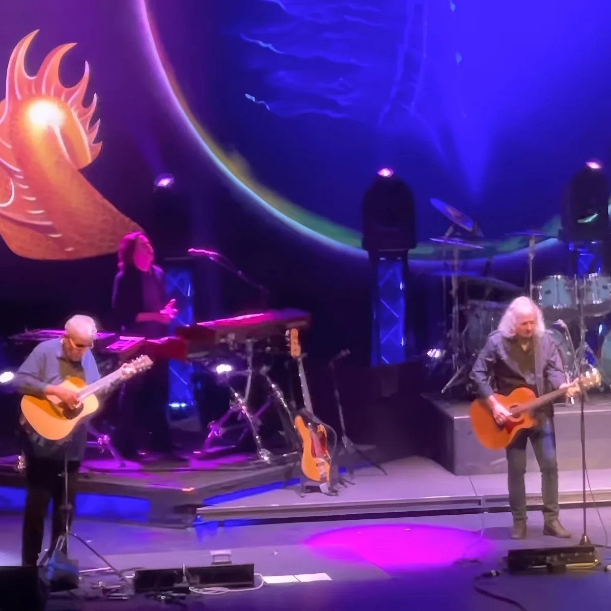A musician plays guitar on stage with a colorful dragon backdrop and other musicians in the background.